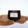 A black container labeled 'KUCHENA SUGAR SCRUB' with 'Monoi' fragrance on a rustic wooden slice.