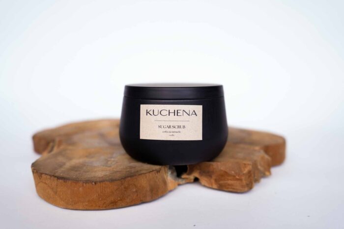 A black container labeled 'KUCHENA SUGAR SCRUB' with 'Monoi' fragrance on a rustic wooden slice.