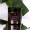 A glass jar of KUCHENA Face Cream with a black lid, labeled as non-comedogenic and suitable for sensitive and acne-prone skin, placed in front of green leaves.