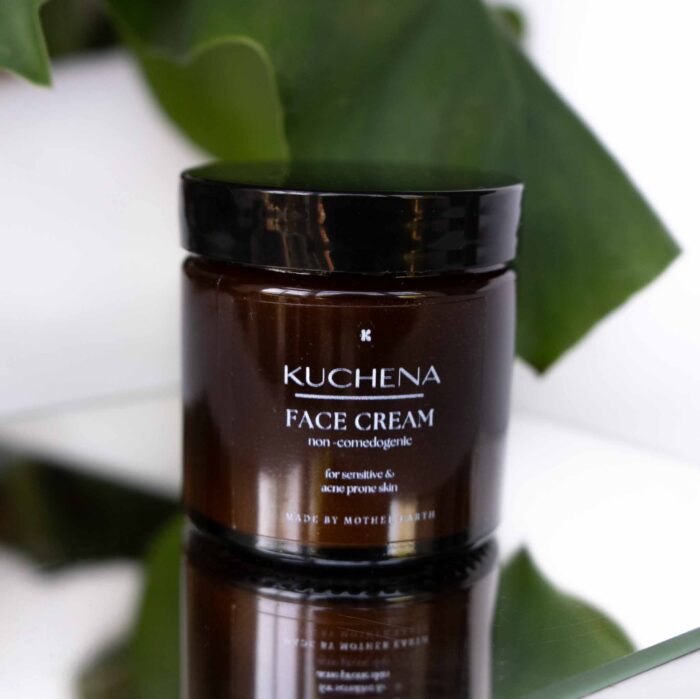 A glass jar of KUCHENA Face Cream with a black lid, labeled as non-comedogenic and suitable for sensitive and acne-prone skin, placed in front of green leaves.