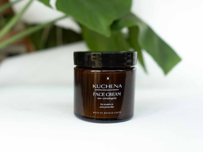 A brown glass jar of Kuchena Face Cream is placed on a white surface with green leaves in the background. The label on the jar indicates it is non-comedogenic and suitable for sensitive and acne-prone skin.