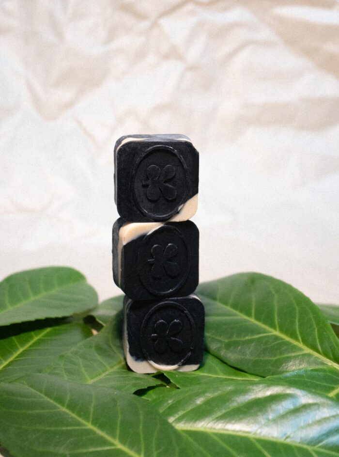 Actieve houtskool en lemongrass zeep - Cube trio en stapel van drie zwarte zeepblokjes met een ingegraveerd klaverblad, gepresenteerd op groene bladeren tegen een lichte, neutrale achtergrond. De zeep is gemaakt van natuurlijke ingrediënten, waaronder actieve houtskool en lemongrass.